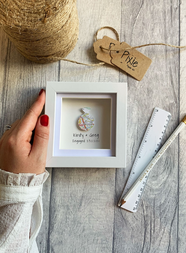 Pu&ograve; includere: Una cornice bianca con una mappa di una citt&agrave; a forma di anello. Il testo "Kirsty & Greg Engaged 23.11.2021" &egrave; sotto l'anello.