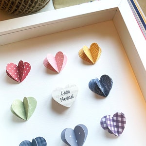 May include: A white frame with a white background and nine paper hearts. The hearts are in various colors and patterns, including red with white polka dots, pink with white stripes, yellow with white polka dots, green with white polka dots, blue with white polka dots, blue with white stripes, purple with white gingham, and a white heart with music notes and the text "Cadoo Medical".