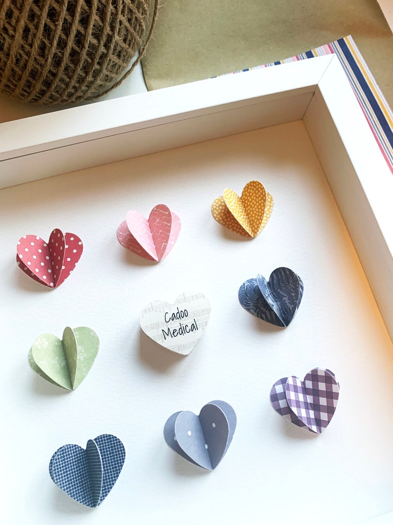 May include: A white frame with a white background and nine paper hearts. The hearts are in various colors and patterns, including red with white polka dots, pink with white stripes, yellow with white polka dots, green with white polka dots, blue with white polka dots, blue with white stripes, purple with white gingham, and a white heart with music notes and the text "Cadoo Medical".