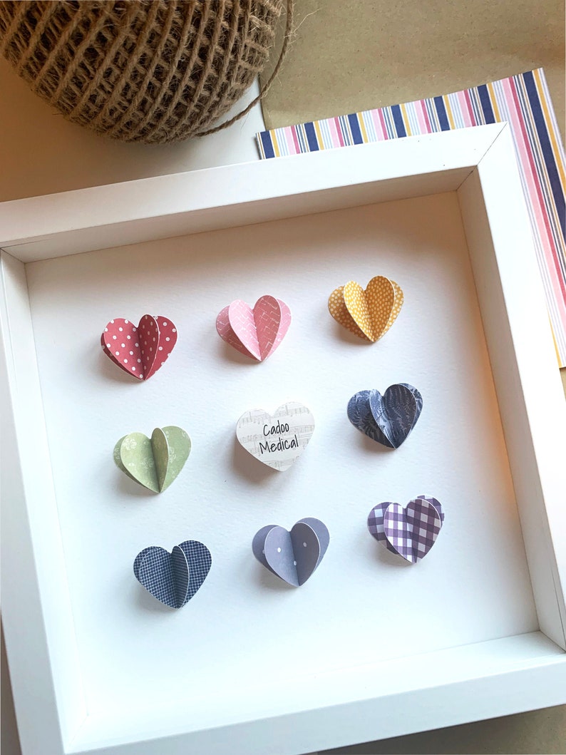May include: A white frame with a white background containing nine paper hearts. The hearts are decorated with different patterns, including polka dots, plaid, and floral designs. The text "Cadoo Medical" is written on one of the hearts.