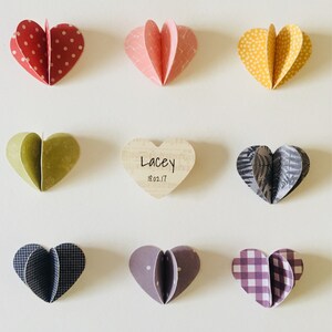 May include: Nine paper hearts with different patterns and colors, including red, yellow, green, pink, purple, and black. The hearts are arranged in a grid pattern on a white background. A wooden heart with the text "Lacey 18.02.17" is in the center.
