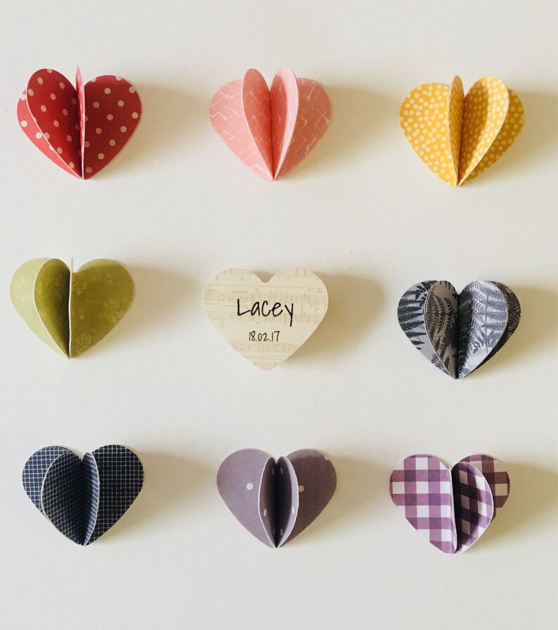 May include: Nine paper hearts with different patterns and colors, including red, yellow, green, pink, purple, and black. The hearts are arranged in a grid pattern on a white background. A wooden heart with the text "Lacey 18.02.17" is in the center.
