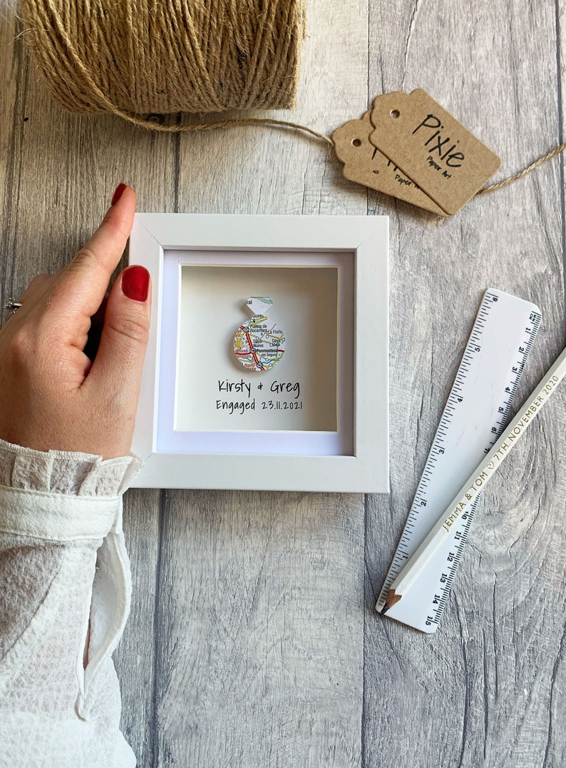 Pu&ograve; includere: Una cornice bianca con una mappa di una citt&agrave; piegata a forma di anello. Il testo "Kirsty & Greg Engaged 23.11.2021" &egrave; scritto sotto l'anello.