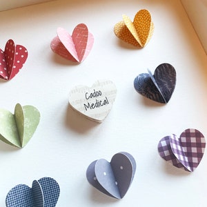 May include: A white frame with a heart-shaped wooden plaque that says "Cadoo Medical" surrounded by nine paper hearts in various colors and patterns.