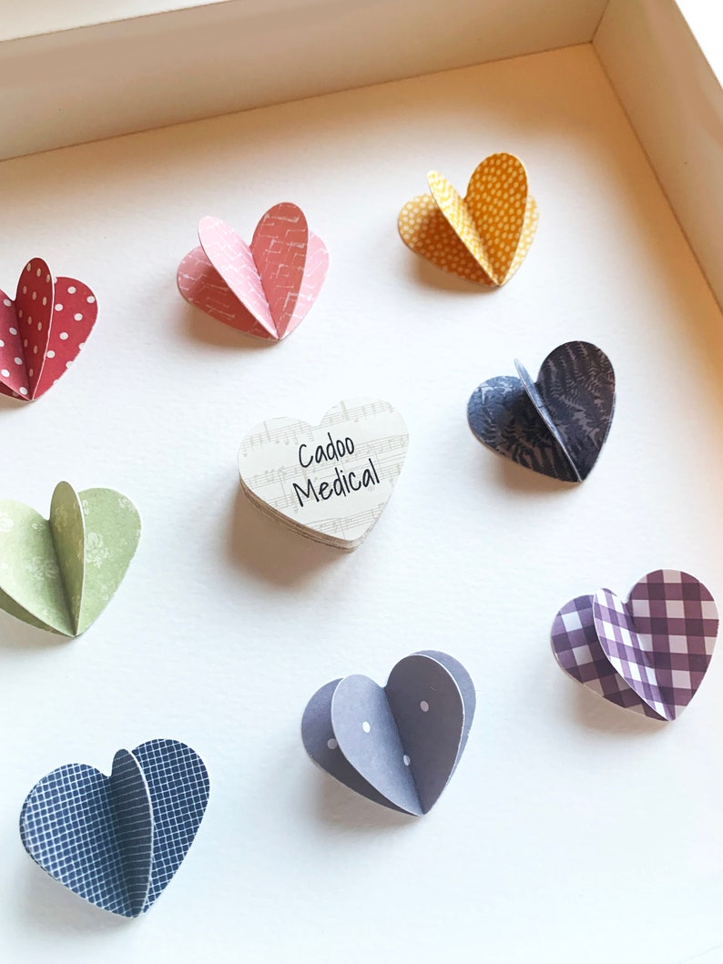 May include: A white frame with a heart-shaped wooden plaque that says "Cadoo Medical" surrounded by nine paper hearts in various colors and patterns.