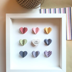 May include: White frame with nine paper hearts in various patterns and colors. The center heart is wooden and says "Cadoo Medical".
