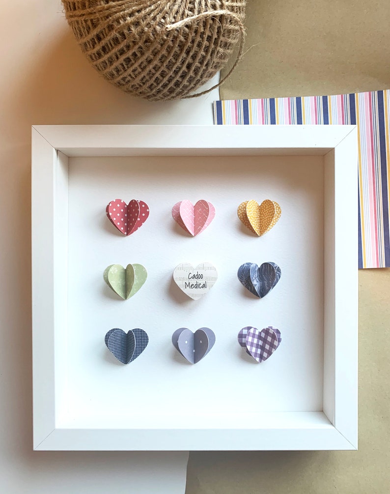 May include: White frame with nine paper hearts in various patterns and colors. The center heart is wooden and says "Cadoo Medical".