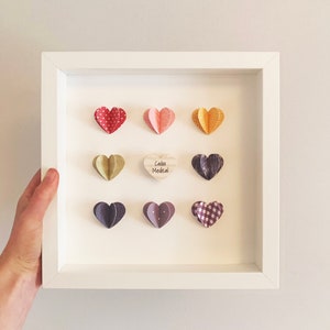 May include: A white frame with a white mat and nine paper hearts in various colors and patterns. The hearts are arranged in a grid pattern. The center heart is a wooden heart with the text "Caddo Medical" written on it.