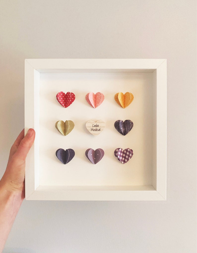 May include: A white frame with a white mat and nine paper hearts in various colors and patterns. The hearts are arranged in a grid pattern. The center heart is a wooden heart with the text "Caddo Medical" written on it.
