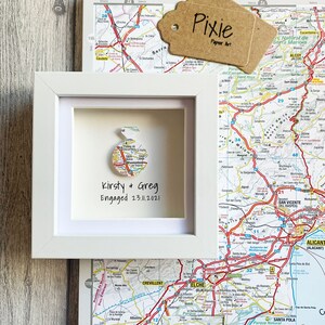Pu&ograve; includere: Una cornice bianca con una mappa della Spagna al suo interno. La mappa &egrave; ritagliata a forma di cuore e il testo "Kirsty & Greg Engaged 23.11.2021" &egrave; scritto sotto la mappa.