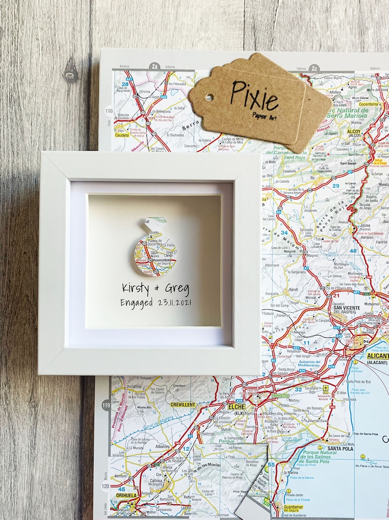Pu&ograve; includere: Una cornice bianca con una mappa della Spagna al suo interno. La mappa &egrave; ritagliata a forma di cuore e il testo "Kirsty & Greg Engaged 23.11.2021" &egrave; scritto sotto la mappa.