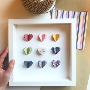 May include: A white frame with a white mat and nine paper hearts inside. The hearts are in various colors and patterns, including polka dots, plaid, and solid colors. The center heart is a wooden heart with the text "Cacao Medical" written on it.
