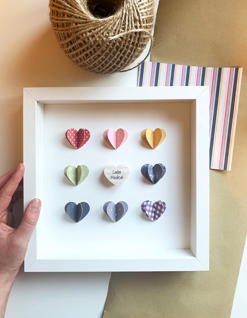 May include: A white frame with a white mat and nine paper hearts inside. The hearts are in various colors and patterns, including polka dots, plaid, and solid colors. The center heart is a wooden heart with the text "Cacao Medical" written on it.