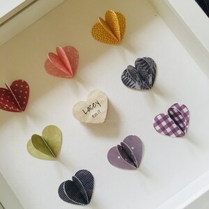 May include: A white frame with a wooden heart in the center that says "Lacey 18.02.17".  Surrounding the wooden heart are eight paper hearts in various colors and patterns, including red, pink, yellow, green, purple, black, and white.