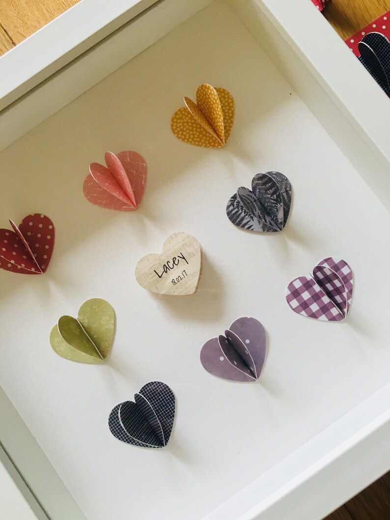 May include: A white frame with a wooden heart in the center that says "Lacey 18.02.17".  Surrounding the wooden heart are eight paper hearts in various colors and patterns, including red, pink, yellow, green, purple, black, and white.