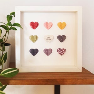 May include: White frame with a white background and nine paper hearts in various colors and patterns. The center heart has the text "Cabo Medical" on it.