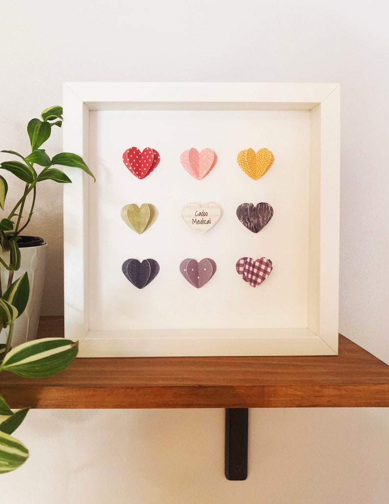 May include: White frame with a white background and nine paper hearts in various colors and patterns. The center heart has the text "Cabo Medical" on it.