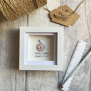 Pu&ograve; includere: Una cornice bianca con un passepartout bianco e una mappa di una citt&agrave; all'interno. La mappa &egrave; piegata a forma di anello ed &egrave; circondata da un bordo bianco. Il testo "Kirsty & Greg Engaged 23.11.2021" &egrave; stampato sotto la mappa.