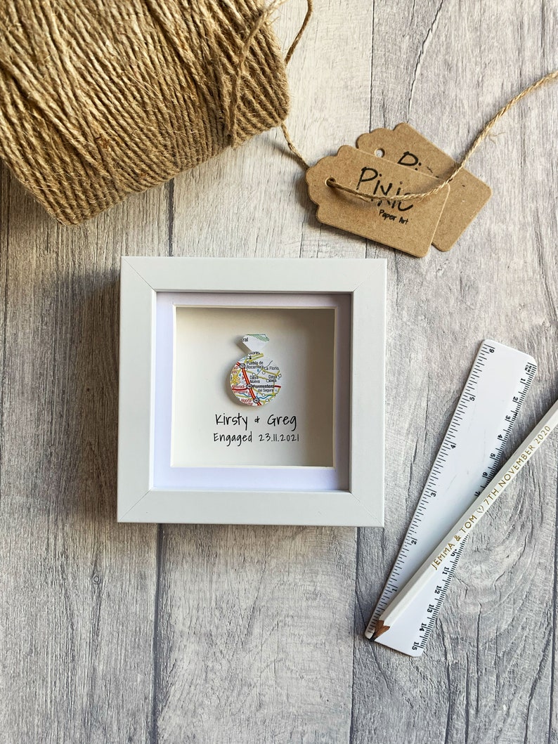 Pu&ograve; includere: Una cornice bianca con un passepartout bianco e una mappa di una citt&agrave; all'interno. La mappa &egrave; piegata a forma di anello ed &egrave; circondata da un bordo bianco. Il testo "Kirsty & Greg Engaged 23.11.2021" &egrave; stampato sotto la mappa.