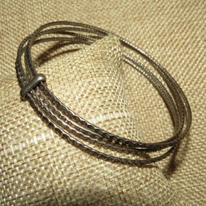 May include: A silver-coloured bracelet featuring four textured bands, joined by a small clasp. The bracelet is displayed on a textured, beige surface, highlighting its detailed design and metallic lustre. A piece of jewellery.