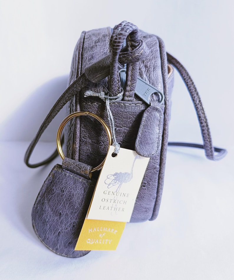 Pode incluir: Uma mala a tiracolo cinzenta com acabamento em pele de avestruz texturada, com um anel de metal dourado e uma etiqueta creme com uma ilustra&ccedil;&atilde;o de avestruz e o texto Genuine Ostrich Leather e Hallmark of Quality.