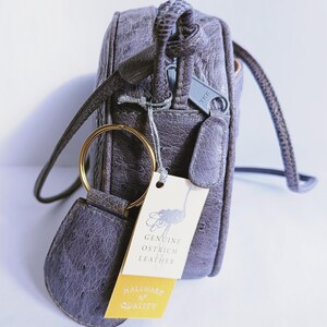Pode incluir: Uma mala a tiracolo cinzenta com acabamento em pele de avestruz texturada, com um anel de metal dourado e uma etiqueta creme com uma ilustra&ccedil;&atilde;o de avestruz e o texto Genuine Ostrich Leather e Hallmark of Quality.