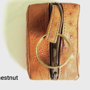 May include: A rectangular chestnut brown leather key pouch featuring a textured surface with small circular indentations, a central metal zip closure, and a large circular gold-toned metal key ring attached to the top tab.