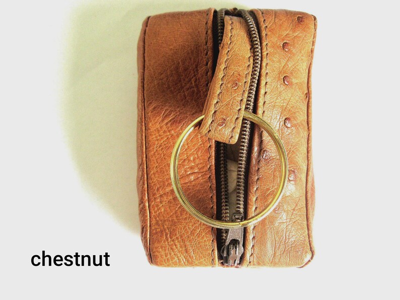 May include: A rectangular chestnut brown leather key pouch featuring a textured surface with small circular indentations, a central metal zip closure, and a large circular gold-toned metal key ring attached to the top tab.