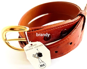 May include: A brandy-coloured ostrich leather belt featuring a polished gold-tone metal buckle. A white tag attached to the belt displays the text "GENUINE OSTRICH LEATHER," an illustration of an ostrich, and the letters "KKLK" inside a circle.
