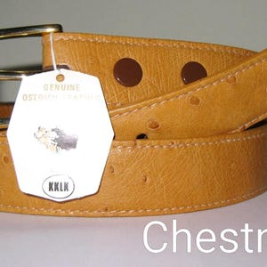 May include: A tan belt with a textured surface and a gold-toned metal buckle. A white tag attached to the belt reads Genuine Ostrich Leather, features a small graphic, and includes the text KKLK. The word Chestnut is printed below.