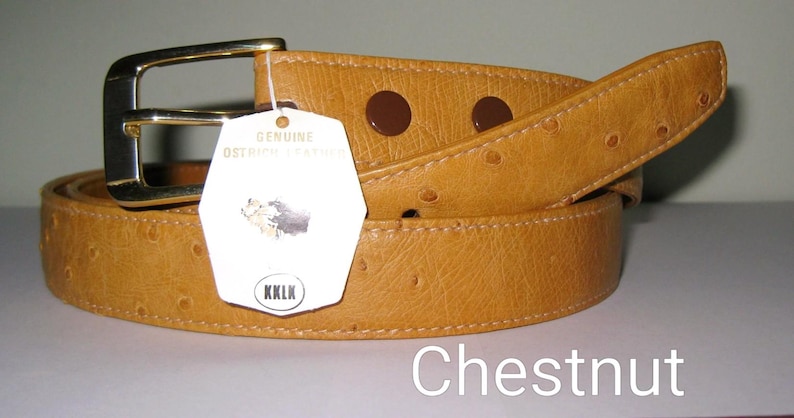May include: A tan belt with a textured surface and a gold-toned metal buckle. A white tag attached to the belt reads Genuine Ostrich Leather, features a small graphic, and includes the text KKLK. The word Chestnut is printed below.
