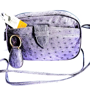 Pode incluir: Uma bolsa tiracolo de couro roxo com textura de avestruz, al&ccedil;a longa e fina, bolso frontal com fecho de aba central e um porta-chaves circular dourado. Uma pequena etiqueta branca e amarela com um log&oacute;tipo est&aacute; inserida no bolso frontal.