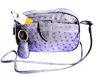 May include: A purple ostrich-textured leather crossbody bag with a long thin strap, a front pocket with a central tab closure, and a gold-toned circular key ring attachment. A small white and yellow tag with a graphic logo is tucked into the pocket.