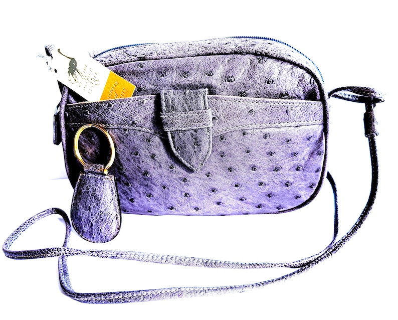 Pode incluir: Uma bolsa tiracolo de couro roxo com textura de avestruz, al&ccedil;a longa e fina, bolso frontal com fecho de aba central e um porta-chaves circular dourado. Uma pequena etiqueta branca e amarela com um log&oacute;tipo est&aacute; inserida no bolso frontal.