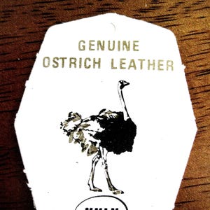 May include: A white, shield-shaped tag featuring the text GENUINE OSTRICH LEATHER in gold lettering. Below the text is a black and white illustration of an ostrich. At the bottom, the letters KKLK are printed inside a black oval outline.