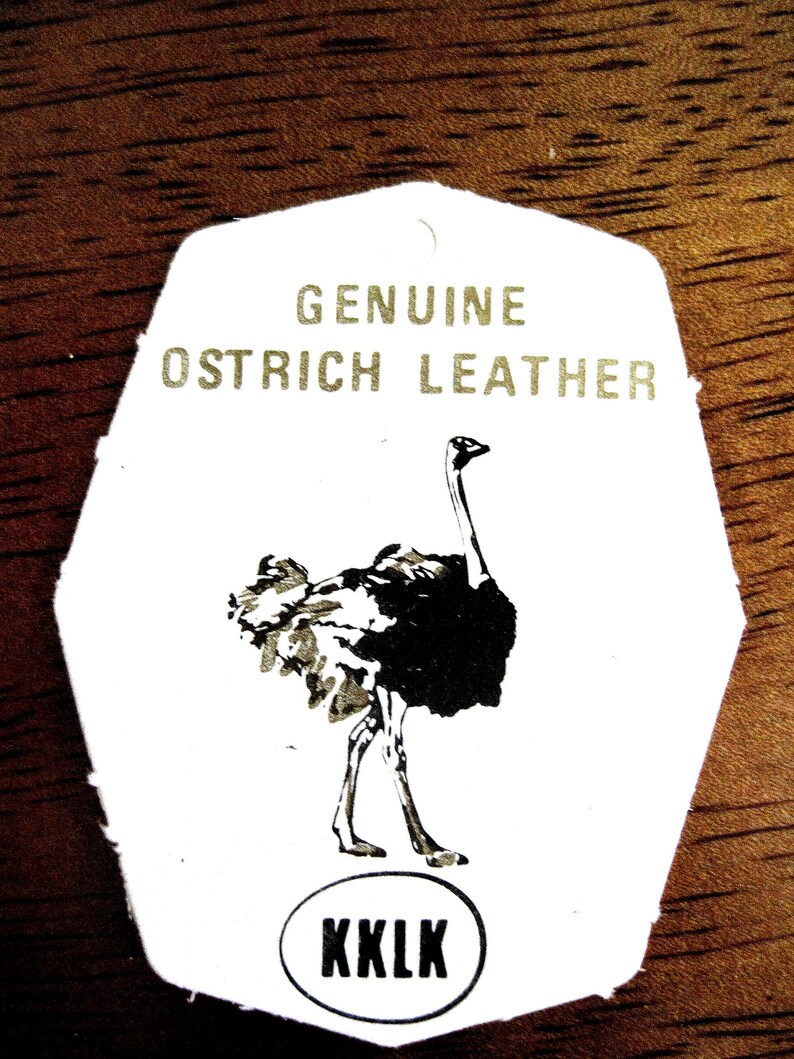 May include: A white, shield-shaped tag featuring the text GENUINE OSTRICH LEATHER in gold lettering. Below the text is a black and white illustration of an ostrich. At the bottom, the letters KKLK are printed inside a black oval outline.