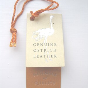 Pode incluir: Uma etiqueta retangular de cor creme com um gr&aacute;fico de avestruz prateado e o texto GENUINE OSTRICH LEATHER, presa a uma etiqueta castanha mais pequena com o texto prateado HALLMARK of QUALITY, tudo suspenso por um cord&atilde;o laranja entran&ccedil;ado.