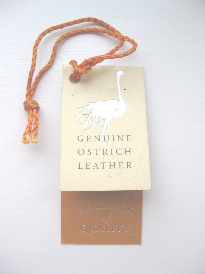 Pode incluir: Uma etiqueta retangular de cor creme com um gr&aacute;fico de avestruz prateado e o texto GENUINE OSTRICH LEATHER, presa a uma etiqueta castanha mais pequena com o texto prateado HALLMARK of QUALITY, tudo suspenso por um cord&atilde;o laranja entran&ccedil;ado.