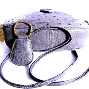 Pode incluir: Uma bolsa transversal cinza com uma superf&iacute;cie texturizada com padr&atilde;o de penas. A bolsa possui um chaveiro de couro cinza correspondente preso a um anel de metal dourado. Uma al&ccedil;a longa e fina se estende das laterais da bolsa arredondada.