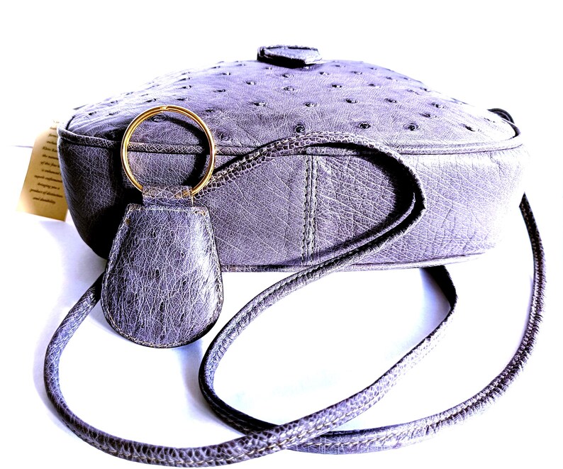 Pode incluir: Uma bolsa transversal cinza com uma superf&iacute;cie texturizada com padr&atilde;o de penas. A bolsa possui um chaveiro de couro cinza correspondente preso a um anel de metal dourado. Uma al&ccedil;a longa e fina se estende das laterais da bolsa arredondada.