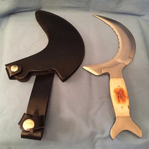 Könnte beinhalten: Ein Messer mit gebogener Klinge, Knochengriff und schwarzer Lederscheide. Die Klinge hat eine gezahnte Kante und eine polierte Oberfläche. Der Griff ist hellbraun mit einem rötlich-braunen Fleck. Die Scheide hat einen Riemen mit Druckknopfverschluss.
