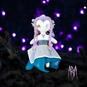 Puede incluir: Una muñeca hecha a mano con cabello morado, un vestido blanco y una falda azul. La muñeca tiene una cara blanca con ojos azules pintados y una nariz dorada. La muñeca lleva un cinturón dorado y un collar con un diseño turquesa. La muñeca está de pie sobre una superficie oscura con un fondo morado. La palabra "ARIA" está escrita en rosa en la superficie.