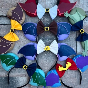 May include: A set of ten colorful mouse ears with various character-themed designs, including a purple and blue pair with a red crown, a green and blue pair with a green dragon, and a brown and yellow pair with a yellow bow.