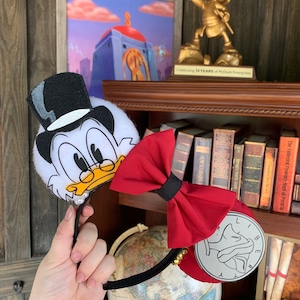 Op de afbeelding: Rode en zwarte strik Minnie Mouse oren met een wit en zwart pluche Scrooge McDuck hoofd met een hoge hoed. De oren hebben een zilveren muntbedel met de tekst "Liberty" erop.