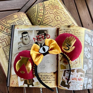 May include: A pair of red and gold Mickey Mouse ears with a gold hand and eye design. The ears are lying on top of an open book with illustrations and text from the show Gravity Falls.