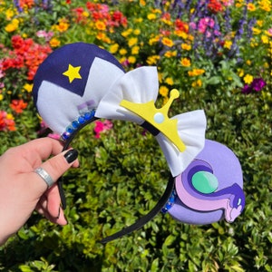 May include: Purple and blue mouse ears with a white bow and a yellow crown. The ears are decorated with blue rhinestones.