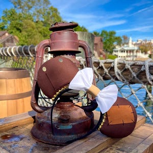 Puede incluir: Orejas de ratón marrones y tostadas con un lazo blanco. Las orejas tienen un ribete de cuentas doradas y un parche marrón en una oreja. Las orejas están sobre una superficie de madera delante de una linterna oxidada.