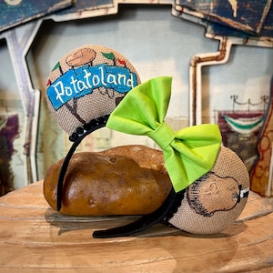 May include: A pair of brown burlap Minnie Mouse ears with a green bow. One ear features a potato character and the text "PotatoLand". The other ear features a potato character with a mustache.