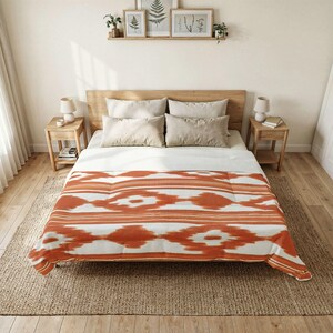 Könnte beinhalten: Ein Bett mit einer weißen Bettdecke mit wiederkehrendem, rostorangem Ikat-Muster, vier beigen Kissen und einem hölzernen Kopfteil. Das Bett steht auf einem strukturierten Teppich in einem Raum mit zwei Nachttischen aus Holz und botanischer Wandkunst.