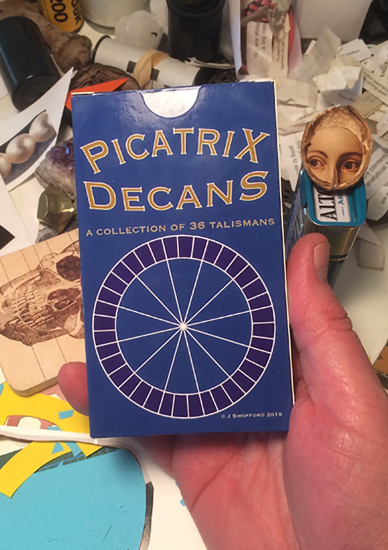 May include: A blue and gold deck of cards called "Picatrix Decans: A Collection of 36 Talismans". The deck features a circular design with a white star in the center and a purple ring around the edge.